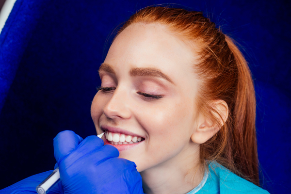 dentist works on tooth crown in dental laboratory ultraviolet painting teeth with mineral and calcium cream to young red-haired ginger woman